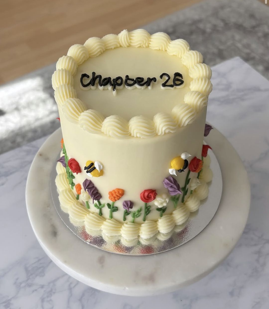 Tall cream vintage cake with shell-piped borders, “Chapter 26” written on top and hand-piped wildflowers and bees around the side