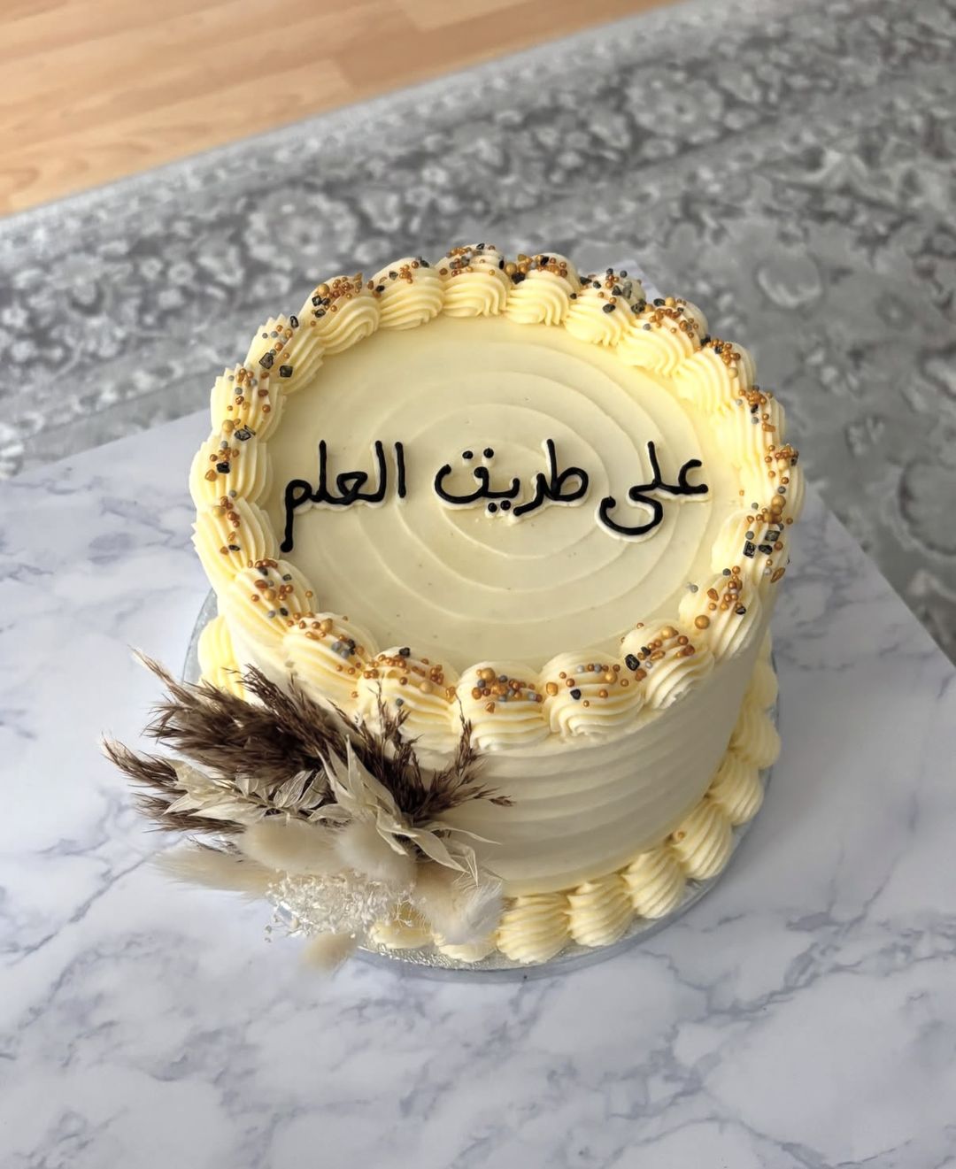 Cream cake with gold-sprinkled piping, Arabic lettering on top and dried pampas flowers on the side (photo 2 of 3)