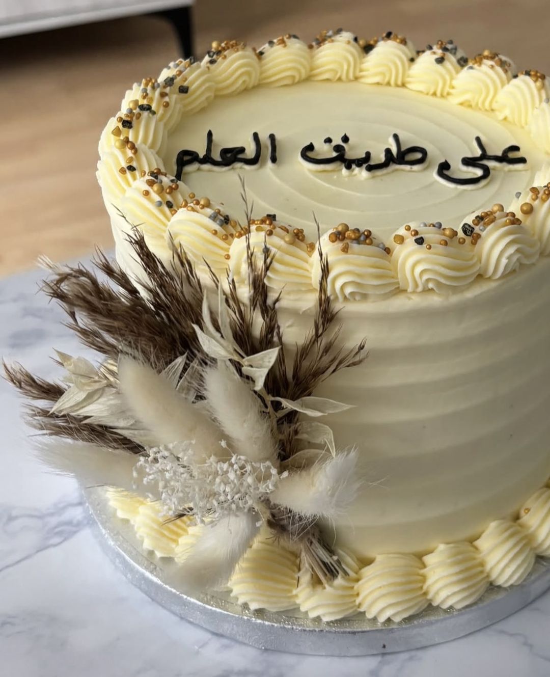 Cream cake with gold-sprinkled piping, Arabic lettering on top and dried pampas flowers on the side (photo 3 of 3)